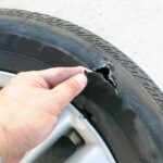 A hand examines a damaged tire with a visible tear and wire exposure.